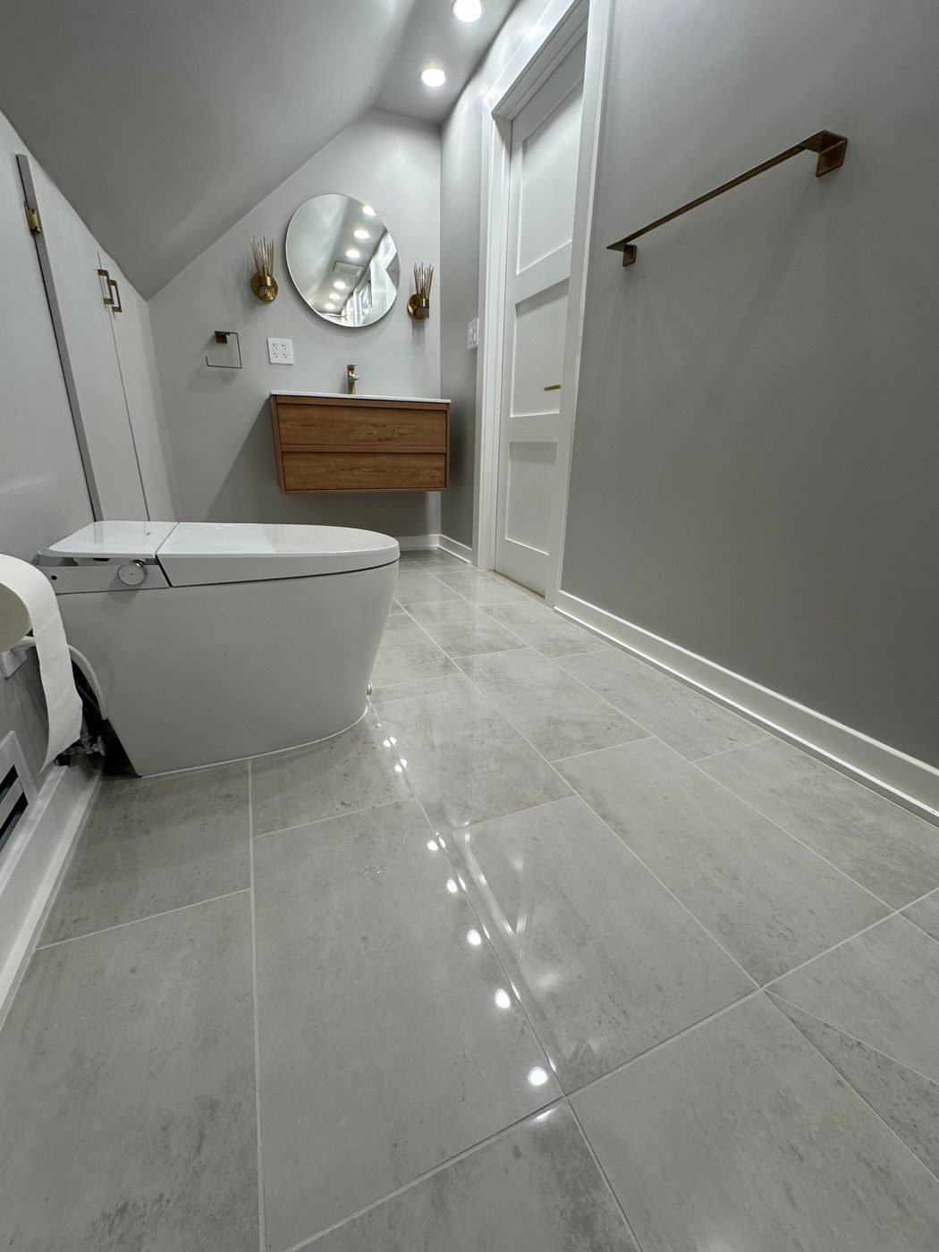 Attic bathroom with a floating wood vanity, round mirror, brass sconces and large gray porcelain floor tile