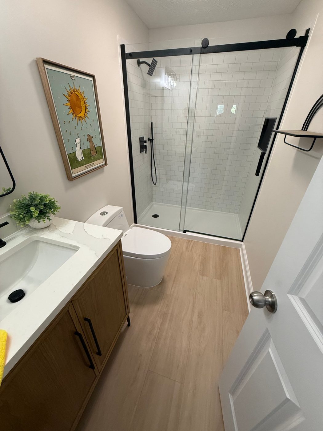 Bathroom with a white subway tile shower behind a black sliding glass door, oak vanity with quartz top and wood-look floor, built by DRUX Tech
