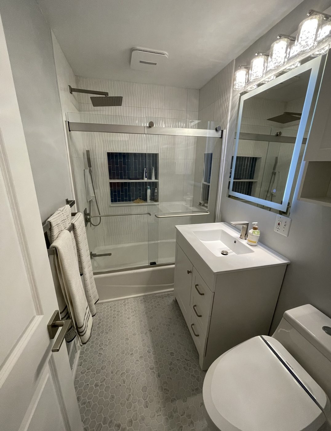 Bathroom with white wave-pattern tile around the tub, a blue tile niche, sliding glass doors, lighted mirror and hex tile floor