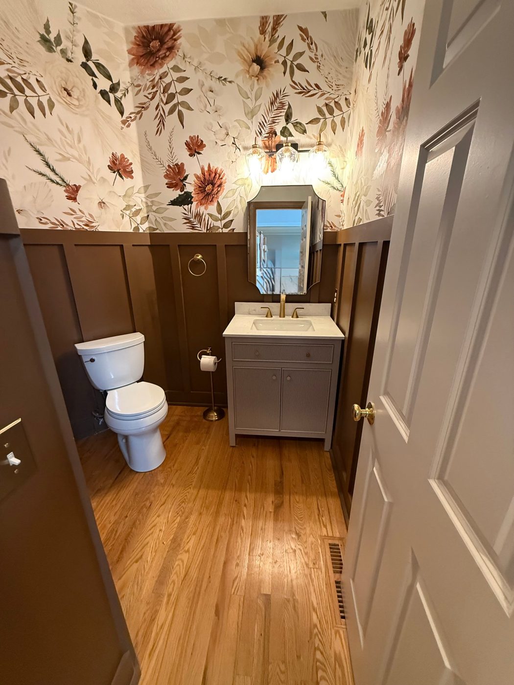 Powder room with floral wallpaper above brown board-and-batten wainscoting, gray vanity and brass fixtures